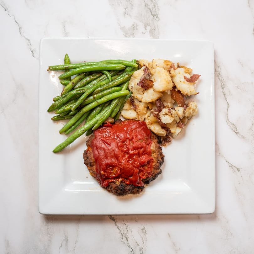 Meatloaf with red sauce, green beans, and roasted potatoes on a white square plate.