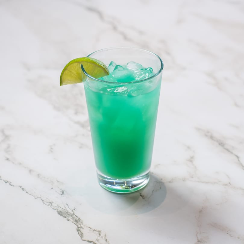 A vibrant blue-green cocktail with ice and a lime wedge on a white marble surface.