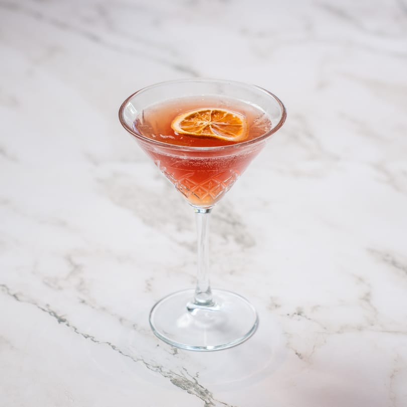 A pink cocktail in a crystal martini glass, garnished with a dried orange slice on a marble surface.